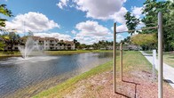This single-floor property is situated within a well-established tropical residential community characterized by its scenic waterfront setting. The exterior grounds are a standout feature, showcasing a tranquil lake that reflects the open Florida sky, lined with lush green lawns and towering palm trees that lend the surroundings a resort-like ambiance. An outdoor fitness station is conveniently positioned along the water's edge, encouraging an active lifestyle amid natural beauty. The community itself features multi-story residential buildings with covered balconies, tile roofing, and mature landscaping, all contributing to a cohesive and appealing aesthetic. The grounds are thoughtfully maintained, with manicured grass and natural ground cover framing the lakefront walkway. The overall atmosphere is peaceful and bright, benefiting from abundant natural sunlight and open skies typical of South Florida's climate. This property offers residents direct access to a picturesque outdoor environment that seamlessly blends recreational amenities with natural surroundings, making it an inviting place to enjoy the outdoors year-round.