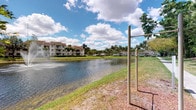 This single-floor property is situated within a well-established tropical residential community characterized by its scenic waterfront setting. The exterior grounds are a standout feature, showcasing a tranquil lake that reflects the open Florida sky, lined with lush green lawns and towering palm trees that lend the surroundings a resort-like ambiance. An outdoor fitness station is conveniently positioned along the water's edge, encouraging an active lifestyle amid natural beauty. The community itself features multi-story residential buildings with covered balconies, tile roofing, and mature landscaping, all contributing to a cohesive and appealing aesthetic. The grounds are thoughtfully maintained, with manicured grass and natural ground cover framing the lakefront walkway. The overall atmosphere is peaceful and bright, benefiting from abundant natural sunlight and open skies typical of South Florida's climate. This property offers residents direct access to a picturesque outdoor environment that seamlessly blends recreational amenities with natural surroundings, making it an inviting place to enjoy the outdoors year-round.