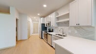 This single-floor unit presents a cohesive, modern aesthetic throughout, anchored by light hardwood flooring that flows seamlessly from room to room. A hallway with natural-toned wood doors leads into the main living and kitchen areas, creating a welcoming sense of arrival.

The kitchen is a standout feature, outfitted with white shaker-style cabinetry, open shelving, and a striking marble-look tile backsplash. White quartz countertops provide ample workspace, while a full suite of stainless steel appliances — including a refrigerator, electric range, microwave, and dishwasher — ensures full functionality. A window above the sink area allows natural light to filter in, framing views of surrounding greenery.

The open living area is bright and airy, with recessed lighting and a wall-mounted mini-split unit providing climate control. A sliding glass door leads directly to a private balcony, offering outdoor views of lush trees and enhancing the connection to nature. A baseboard heater runs along the exterior wall, supplementing the heating system.

The bedroom is a calm, neutral space with a closet rod and clean walls. The bathroom features a grey vanity with white countertop, a large mirror, and light flooring, maintaining the unit's clean, contemporary palette throughout.