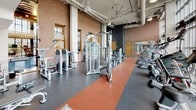 Located at 77 H St NW in Washington, DC, this fitness center offers a comprehensive and thoughtfully designed workout environment across a single floor. The space is defined by dramatic floor-to-ceiling curtain-wall windows that line the perimeter, flooding the interior with natural light and providing open views of the surrounding cityscape.

The cardio zone is positioned directly along the windowed facade, housing a full row of treadmills, ellipticals, and stair climbers — allowing members to exercise while taking in views of the DC skyline. Overhead, large industrial ceiling fans circulate air throughout the space, complementing the exposed ductwork and concrete structural elements that give the facility a modern, industrial aesthetic.

The strength training area features an extensive selection of cable machines, selectorized weight stations, and free weights, including a full dumbbell rack. Warm brick walls and a distinctive orange rubber runway flooring create visual contrast against the gray rubberized workout surfaces. Mirrors are strategically placed to enhance sightlines throughout.

A dedicated functional training and stretching zone is equipped with stability balls, reformer equipment, and open floor space. A separate corridor area with large windows offers additional workout stations and shelving for personal items. The overall ambiance is energetic yet functional, providing a well-rounded fitness experience in the heart of Washington, DC.