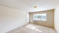 Located in Spokane Valley, WA, this one-bedroom, one-bathroom unit offers a practical and efficiently designed floor plan across a single level. The living room is bright and welcoming, featuring plush carpeting, a large window that frames outdoor greenery, a private entry door, and a wall-mounted A/C unit. The open-concept kitchen flows seamlessly into the living area and is appointed with warm wood cabinetry, light countertops, black appliances including an electric range and microwave, and a peninsula with pendant lighting overhead — ideal for casual dining or meal prep. The bedroom is a comfortable, carpeted retreat with a window overlooking the surrounding landscape and a closet for storage. The full bathroom includes a vanity with wood cabinetry, tile flooring, a baseboard heater, and a shower/tub combination. A dedicated laundry area with a full-size washer and dryer and an overhead wire shelf adds everyday convenience. Neutral tones and soft carpeting throughout lend the space a clean, move-in-ready aesthetic suited for comfortable everyday living.