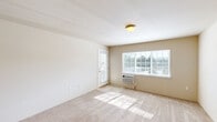 Located in Spokane Valley, WA, this one-bedroom, one-bathroom unit offers a practical and efficiently designed floor plan across a single level. The living room is bright and welcoming, featuring plush carpeting, a large window that frames outdoor greenery, a private entry door, and a wall-mounted A/C unit. The open-concept kitchen flows seamlessly into the living area and is appointed with warm wood cabinetry, light countertops, black appliances including an electric range and microwave, and a peninsula with pendant lighting overhead — ideal for casual dining or meal prep. The bedroom is a comfortable, carpeted retreat with a window overlooking the surrounding landscape and a closet for storage. The full bathroom includes a vanity with wood cabinetry, tile flooring, a baseboard heater, and a shower/tub combination. A dedicated laundry area with a full-size washer and dryer and an overhead wire shelf adds everyday convenience. Neutral tones and soft carpeting throughout lend the space a clean, move-in-ready aesthetic suited for comfortable everyday living.