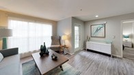 The Willow is a thoughtfully designed one-bedroom, one-bathroom single-floor residence that balances comfort and modern style. The open-concept living area is bathed in natural light from large sliding glass doors and windows dressed with sheer curtains, creating a bright and welcoming atmosphere. Wood-style plank flooring runs throughout the main living spaces, adding warmth and a contemporary feel.

The kitchen is well-appointed with dark espresso cabinetry, granite-style countertops, black appliances including a range and refrigerator, and a peninsula that doubles as a breakfast bar — seamlessly connecting the kitchen to the living and dining area for easy entertaining.

The bedroom offers plush carpeting, a ceiling fan with light fixture, and generous window coverage that allows for ample natural light while maintaining privacy. The space comfortably accommodates a full-sized bed and additional furnishings.

The bathroom features a full tub and shower combination with white tile surround, a granite-topped vanity with an undermount sink, wood-tone cabinetry, and wood-style flooring that ties in with the rest of the unit. Additional storage is provided by a tall built-in cabinet.

Overall, the Willow presents a clean, neutral palette with modern touches, making it a functional and stylish living space.