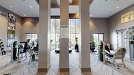 This well-appointed fitness center in Gainesville, Florida, offers a generously sized, single-floor layout divided into two dedicated exercise areas. The space makes a striking first impression with soaring ceilings, bold architectural columns, and floor-to-ceiling windows that flood the interior with natural light and frame views of lush palm-lined greenery outdoors.

The main exercise area is comprehensively equipped with cardio machines, including treadmills, ellipticals, and stationary bikes, all positioned along the expansive window wall to take full advantage of the outdoor scenery. A dedicated strength-training zone features cable machines, weight racks with free weights, benches, and a variety of resistance equipment to support a complete workout routine.

A separate room, accessible via a sliding barn-style door, provides additional workout space with mirrored walls and hardwood-style flooring, offering an ideal environment for stretching, functional training, or group exercise. Decorative artwork with vibrant, mid-century modern-inspired prints and motivational framed photography adds personality and energy to the space.

The interior finishes are modern and polished, featuring herringbone-patterned carpet, recessed lighting, pendant fixtures, and acoustic ceiling panels. The overall ambiance is bright, open, and motivating, creating an inviting environment for residents to pursue their fitness goals.