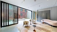 Located at 598 W Main St in Charlottesville, Virginia, this single-floor lounge space presents a refined, gallery-like environment defined by clean architectural lines and a carefully considered aesthetic. The interior is characterized by white walls, track lighting, and a neutral palette of cream, blush, and warm taupe tones that create a calm, sophisticated ambiance. Curved, sculptural seating arrangements anchored by layered area rugs lend the space an organic softness, while large-scale fine art pieces displayed on the walls reinforce its gallery sensibility.

Floor-to-ceiling glass sliding walls dissolve the boundary between indoors and outdoors, opening directly onto a well-designed courtyard. The courtyard features curved planting beds with young trees and ground cover, smooth concrete paving, linear slatted benches, and a Corten steel-framed fireplace set against an exposed brick backdrop, creating a compelling outdoor gathering area. A separate landscaped passage along the building's exterior adds another layer of greenery and texture.

Translucent drapery panels serve as elegant room dividers within the interior, adding depth without sacrificing the open, airy quality of the space. The seamless integration of indoor lounge and outdoor courtyard, combined with thoughtful art curation and contemporary furnishings, gives this space a distinctive character suited for elevated social or professional use.