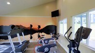 This single-floor fitness center is thoughtfully designed to inspire and energize those who use it. The space is immediately defined by a dramatic, full-wall mural depicting the silhouettes of hikers against a vivid sunset landscape reflected in still water — a striking visual centerpiece that brings an adventurous, motivating atmosphere to every workout. The warm, golden tones of the mural are complemented by light-colored walls and neutral flooring, creating a bright and welcoming environment. A wall-mounted flat-screen television is positioned at an ideal viewing height, offering entertainment during exercise sessions. The fitness area is outfitted with multiple LifeCycle stationary bikes and additional cardio equipment, including what appears to be an elliptical machine and a rowing or step machine, providing a well-rounded selection of cardiovascular workout options. Large windows along one side of the room allow abundant natural light to flood the space and offer views of the surrounding greenery outdoors, enhancing the open and airy feel of the room. The combination of purposeful equipment layout, inspiring décor, and natural light makes this fitness center a functional and visually engaging space suited for a range of cardio-focused exercise routines.