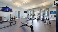 Located at 8890 Ransley Station Blvd in Pensacola, Florida, this fitness center spans a single floor and is divided into two dedicated exercise rooms, offering a comprehensive workout environment for residents or members. The space is finished with durable speckled rubber flooring throughout, providing both safety and functionality, while soft blue-gray walls lend a calm, modern aesthetic.

The main exercise room is generously proportioned and filled with natural light streaming through large, black-framed windows that offer views of the surrounding landscaped grounds. It is equipped with a full array of cardio machines — including treadmills, ellipticals, and a stair climber — alongside free weights, a weight bench, and a power rack. A large wall-mounted mirror enhances the sense of space, and overhead track and recessed lighting ensure excellent visibility. A wall-mounted TV adds to the convenience of the space.

The second exercise room serves as a dedicated functional and low-impact training area. It features tall ceilings and floor-to-ceiling windows that flood the room with natural light and outdoor views. Equipment includes stability balls, resistance bands, foam rollers, a WaterRower rowing machine, and a Peloton cycling bike, making it ideal for stretching, cross-training, or focused cardio sessions. A water dispenser is also conveniently located within the space.