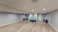 This single-level fitness studio is thoughtfully designed to support a wide range of exercise and movement activities. The space is anchored by a full-length mirrored wall that spans the length of the room, allowing for form checks and creating a sense of expanded depth throughout the studio. The warm hardwood-style flooring provides a durable yet comfortable surface ideal for yoga, dance, pilates, or general training routines. Soft blue-gray walls lend a calm, focused atmosphere to the environment. Recessed lighting in the ceiling provides even, well-distributed illumination across the entire workout area. A centrally located HVAC vent ensures consistent climate control, keeping the space comfortable during intense sessions. The room is equipped with exercise accessories including stability balls and balance trainers, which are neatly arranged along the perimeter. Storage solutions such as a white shelving unit keep equipment organized and accessible. Frosted glass panel doors add a refined touch while maintaining privacy. The overall layout is open and unobstructed, maximizing usable floor space for movement-based activities. This studio offers a clean, professional environment well-suited for personal training, group fitness classes, or private wellness routines.