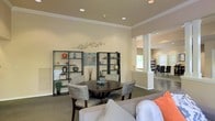 This single-floor space offers an open, well-appointed interior designed for comfort and functionality. The main gathering area features a round dining table surrounded by upholstered chairs, creating an inviting setting for casual meetings or social interaction. Flanking the seating area are two tall, dark-framed open shelving units displaying decorative objects, adding a curated aesthetic to the warm, neutral-toned walls accented by crown molding.

Recessed lighting throughout the ceiling provides consistent, even illumination, while a columned partition elegantly separates the main lounge area from an adjacent space visible in the background, which appears to be a secondary dining or workspace with additional seating. The structural columns add an architectural character to the layout, contributing to the refined yet approachable atmosphere.

Carpeted flooring in the main seating zone transitions toward what appears to be a hard-surface floor in the connecting room. Natural light filters in through windows at the rear, brightening the overall environment. The space is thoughtfully styled with coordinated furnishings, including a neutral sofa with accent cushions, making it well-suited as a community lounge, clubhouse, or shared amenity space.