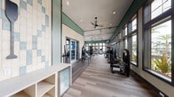 This spacious fitness center is designed to support a comprehensive workout experience across a single open floor. The main exercise area is characterized by soaring ceilings accented with deep teal crown molding and a striking decorative ceiling panel featuring a bold botanical pattern. Light wood-tone plank flooring runs throughout, lending a clean, modern aesthetic. A wall of large, dark-framed windows spans one side of the room, flooding the space with natural light and offering views of palm trees and manicured outdoor landscaping.

The facility is well-stocked with a wide array of equipment, including multiple cable and selectorized strength machines, a free-standing heavy bag, TRX suspension trainers, a functional trainer, and a medicine ball rack. Cardio equipment such as treadmills and step machines from Life Fitness lines the perimeter. Ceiling fans and recessed lighting ensure consistent comfort throughout.

A separate studio area, finished in darker tones with its own tall window overlooking a serene water view, houses spin bikes, a wall-mounted display, a yoga mat, and a colorful dumbbell rack—ideal for individual training sessions. A small built-in refrigeration unit near the entrance adds a practical amenity for post-workout refreshment.