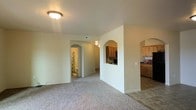 This single-floor space showcases a thoughtfully designed interior highlighted by charming arched doorways that add architectural character throughout. The main living area is finished with neutral-toned carpet, creating a warm and comfortable atmosphere, while tile flooring marks the transition into the kitchen zone. A standout feature is the arched pass-through window connecting the living area to the kitchen, allowing for an open feel and easy interaction between spaces without fully removing the wall partition. The kitchen is equipped with natural wood-toned cabinetry, providing ample storage and a classic, homey aesthetic. A flush-mount ceiling light fixture illuminates the living space, contributing to the clean, understated look of the interior. Arched doorways lead to additional areas, including a bathroom visible in the background, which features a shower with a decorative curtain. The neutral wall tones throughout lend the space a cohesive and versatile canvas. Overall, the layout balances openness and defined zones, making efficient use of the available space across its single level.