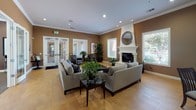 This single-story clubhouse is designed to serve as a versatile gathering space, combining comfort and functionality across several distinct areas. The main lounge features wood-style tile flooring, crown molding, and a striking white fireplace with a decorative round mirror above the mantle. Large windows with slatted blinds frame outdoor views of what appears to be a pool area, filling the room with natural light. Neutral gray sofas and a glass-top coffee table complete the welcoming seating arrangement. French doors connect this space to adjacent rooms, maintaining an open, flowing layout. A dedicated media room offers carpeted floors, rich dark brown walls, a wall-mounted flat-screen TV flanked by black cabinetry, and comfortable upholstered seating arranged for optimal viewing. The billiards room features a full-size pool table centered beneath a trio of pendant lights, surrounded by framed classic movie posters and additional bar-height seating. Throughout the clubhouse, recessed lighting, crown molding, and consistent warm brown wall tones create a cohesive, inviting atmosphere suited for both relaxed socializing and recreational activities.
