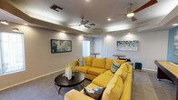 This single-floor lounge space is designed for social gathering and relaxation, combining vibrant color choices with functional furnishings. A striking tray ceiling with recessed lighting and dual ceiling fans serves as the architectural centerpiece, adding dimension and ensuring comfortable airflow throughout the space. The interior is anchored by a large, bold yellow sectional sofa accented with patterned throw pillows, creating an inviting focal point that contrasts effectively with the neutral gray carpeting and warm beige walls.

A vivid teal-blue accent wall on one side adds a dynamic pop of color, complemented by curated abstract artwork displayed throughout the space. A wall-mounted flat-screen television is positioned opposite the sectional, enhancing the room's entertainment functionality. Adjacent to the seating area, a shuffleboard table provides additional recreational options for guests.

Multiple bar-height round tables paired with sleek chrome-legged stools are distributed across the space, offering flexible casual seating arrangements. Vertical blind-covered windows allow natural light to filter in while maintaining privacy. Decorative vases and small potted plants add finishing touches, contributing to the space's welcoming and lively atmosphere.