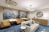Located at 1151 4th St SW in Washington, DC, this thoughtfully designed lounge space combines modern aesthetics with functional comfort. The single-floor layout makes efficient use of its intimate footprint, offering distinct seating zones that cater to both casual socializing and focused gatherings. A richly patterned blue area rug anchors the space, providing a striking foundation throughout. One of the most eye-catching features is the bold geometric accent wall adorned with blue and gold hexagonal tile patterns, lending the space a vibrant, artistic character. Comfortable banquette seating with warm golden upholstery lines this area, paired with white-topped tables ideal for small group settings. The lounge area features a neutral cream sofa, accent chairs in white and cognac, and a sleek coffee table, all bathed in warm light from a modern globe chandelier. A floor-to-ceiling black-framed glass partition wall adds architectural drama while allowing visibility into an adjacent space, enhancing the sense of openness. A wood-front credenza with a round mirror above it completes the refined décor. Recessed lighting and a marble-accented work surface further elevate the polished, contemporary feel of this well-appointed lounge.