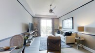 This single-floor space showcases a thoughtfully designed living area that blends modern and rustic elements seamlessly. The room is anchored by light-toned hardwood-style flooring and crisp white walls accented with dark crown molding, creating a clean yet sophisticated contrast. A tray ceiling adds architectural depth to the space, while a contemporary ceiling fan with a sleek metallic finish provides both function and style.

Natural light pours in through a large sliding glass door that opens onto a private balcony, offering outdoor views framed by patterned curtains in soft, muted tones. The living area is comfortably furnished with a neutral-toned sofa adorned with decorative pillows, a woven accent chair, and a patterned area rug that ties the room together. An open media console houses the flat-screen television and decorative accessories, complementing the industrial-rustic aesthetic of the space.

A floor lamp provides warm ambient lighting, enhancing the inviting atmosphere of the room. The overall ambiance is relaxed yet polished, making this space well-suited for both everyday living and entertaining.