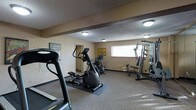 This single-floor fitness space is designed to provide a comprehensive workout environment with a practical and functional layout. The room is outfitted with a variety of exercise equipment, including a treadmill, an elliptical cross-trainer, and a multi-station strength training machine, offering options for both cardiovascular and resistance training. The flooring features durable, cushioned gym-appropriate material that provides support during workouts. A wall-mounted mirror on one side enhances the sense of space and allows users to monitor their form during exercise. Natural light filters in through a window on the far wall, contributing to a bright and energizing atmosphere. The textured ceiling and warm, neutral-toned walls give the room a no-frills, purpose-driven character. Flush-mounted ceiling lights provide even illumination throughout the space. Motivational signage adds a personal touch to the walls, contributing to an encouraging workout environment. A doorway provides access to an adjacent room, adding convenience to the overall layout. This fitness space is well-suited for daily exercise routines and offers a focused environment for achieving health and wellness goals.