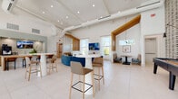 Located on Easton Trace Drive in Sun City Center, Florida, this single-story community lounge is designed with a contemporary aesthetic that balances comfort and functionality. The space features soaring vaulted ceilings with exposed white ductwork and warm wood beam accents, creating an airy, open atmosphere throughout. Large floor-to-ceiling windows and sheer white curtains allow abundant natural light to flood the interior while framing views of the outdoor pool and surrounding Florida landscape.

The main gathering area is anchored by a curved blue sectional sofa and complemented by white pedestal bistro tables with houndstooth-upholstered bar stools, offering flexible seating configurations. A wall-mounted flat-screen television serves as a focal point in this lounge zone. Slatted warm wood columns provide subtle spatial definition without obstructing the open layout.

A dedicated booth seating area features colorful plaid banquettes, a walnut-topped dining table, and a globe pendant light, creating an intimate nook ideal for small gatherings. A compact coffee bar station with built-in cabinetry, a professional beverage machine, a sink, and counter seating offers a convenient refreshment area. Terrazzo flooring unifies the space, enhancing its bright, clean, and welcoming ambiance throughout.