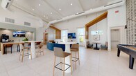 Located on Easton Trace Drive in Sun City Center, Florida, this single-story community lounge is designed with a contemporary aesthetic that balances comfort and functionality. The space features soaring vaulted ceilings with exposed white ductwork and warm wood beam accents, creating an airy, open atmosphere throughout. Large floor-to-ceiling windows and sheer white curtains allow abundant natural light to flood the interior while framing views of the outdoor pool and surrounding Florida landscape.

The main gathering area is anchored by a curved blue sectional sofa and complemented by white pedestal bistro tables with houndstooth-upholstered bar stools, offering flexible seating configurations. A wall-mounted flat-screen television serves as a focal point in this lounge zone. Slatted warm wood columns provide subtle spatial definition without obstructing the open layout.

A dedicated booth seating area features colorful plaid banquettes, a walnut-topped dining table, and a globe pendant light, creating an intimate nook ideal for small gatherings. A compact coffee bar station with built-in cabinetry, a professional beverage machine, a sink, and counter seating offers a convenient refreshment area. Terrazzo flooring unifies the space, enhancing its bright, clean, and welcoming ambiance throughout.