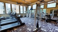 This fitness center offers a comprehensive workout environment designed to accommodate a range of exercise needs. The space is lined with large windows on multiple sides, flooding the interior with natural light and providing views of the surrounding outdoor area, creating an open and energizing atmosphere. The patterned carpet in shades of brown, gold, and grey adds a distinctive visual element to the floor. Cardio equipment, including multiple treadmills, is positioned along the perimeter, while a cable pulley system and various strength-training machines occupy the central area. A wall of mirrors along one side enhances the sense of depth and allows users to monitor their form during workouts. Ceiling fans with warm yellow-toned upper panels are mounted overhead, contributing to air circulation and comfort throughout sessions. A mounted monitor is visible near the cable system, offering entertainment or instructional content. The overall layout is organized and spacious, allowing ample room to move between equipment. The combination of natural light, mirrored walls, and modern fitness equipment makes this a functional and inviting space for daily exercise.