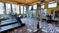 This fitness center offers a comprehensive workout environment designed to accommodate a range of exercise needs. The space is lined with large windows on multiple sides, flooding the interior with natural light and providing views of the surrounding outdoor area, creating an open and energizing atmosphere. The patterned carpet in shades of brown, gold, and grey adds a distinctive visual element to the floor. Cardio equipment, including multiple treadmills, is positioned along the perimeter, while a cable pulley system and various strength-training machines occupy the central area. A wall of mirrors along one side enhances the sense of depth and allows users to monitor their form during workouts. Ceiling fans with warm yellow-toned upper panels are mounted overhead, contributing to air circulation and comfort throughout sessions. A mounted monitor is visible near the cable system, offering entertainment or instructional content. The overall layout is organized and spacious, allowing ample room to move between equipment. The combination of natural light, mirrored walls, and modern fitness equipment makes this a functional and inviting space for daily exercise.