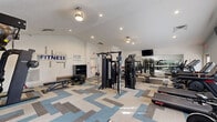 Located at Heron Springs in Stow, Ohio, this well-equipped fitness center offers a comprehensive workout environment spread across a generously sized single-floor layout. The space features vaulted ceilings with recessed lighting that create an open, airy atmosphere, while soft blue-gray walls and a geometric patterned carpet in neutral tones of blue, gray, and beige lend the room a modern, clean aesthetic.

The main workout area is thoughtfully organized to accommodate a full range of exercise disciplines. Cardio equipment includes treadmills, elliptical machines, a rowing machine, and a stationary cycling bike, all positioned near windows that allow natural light to fill the space. Strength training is well-represented with a Nautilus Instinct cable machine, a functional trainer, a weight bench, dumbbells, and additional resistance equipment. Wall-mounted televisions are positioned throughout for entertainment during workouts.

A large floor-to-ceiling mirror spans one wall and is complemented by a ballet barre, creating a dedicated area for stretching, balance training, or dance-style exercise. Motivational fitness-themed wall decals add an energizing touch to the environment. A storage closet and shelving unit provide organizational convenience. Overall, the space delivers a versatile and inviting fitness experience suitable for a wide range of training goals.