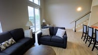 This two-story space offers a well-designed living area characterized by light hardwood flooring and neutral-toned walls that create a clean, understated aesthetic. The living room is a standout feature, boasting floor-to-ceiling windows and a large sliding glass door that floods the space with natural light and provides direct access to an outdoor balcony. The elevated window placement amplifies the sense of vertical space, drawing the eye upward and enhancing the open, airy atmosphere. Outdoor views of greenery are visible through the expansive glazing, adding a connection to the surrounding environment. A staircase with a sleek metal railing is positioned along one wall, leading to the second floor and serving as a subtle architectural detail within the living space. The overall layout appears thoughtfully arranged, balancing functionality with an inviting ambiance. The combination of generous natural light, multi-level design, and access to an outdoor area makes this a versatile and comfortable space suited for both everyday living and entertaining.
