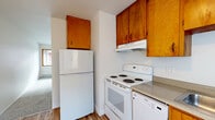 This efficient 2-bedroom, 1-bathroom single-floor unit offers a well-organized layout designed to maximize every square foot. The combined kitchen and living room serves as the heart of the home, where wood-toned cabinetry, white appliances — including a refrigerator, electric range, and dishwasher — and laminate wood-style flooring come together to create a functional and cohesive space. A large wood-trimmed window in the kitchen area allows natural light to flow in, brightening the room throughout the day. The living area features neutral carpet and a clean, open feel with a closet nearby for convenient storage. A connecting hallway leads to the two bedrooms, each finished with soft carpet, wood-trimmed windows, and ceiling fans with integrated lighting. Baseboard heating runs throughout, ensuring consistent warmth. The primary bedroom offers a comfortable footprint with an outlook to the outdoors. The shared bathroom is neatly appointed with a tiled tub-shower combination, a white vanity with a sleek countertop, an oversized mirror, and wood-style flooring. Five closets distributed throughout the unit provide generous storage. The overall aesthetic is clean and neutral, with white walls and warm wood accents lending a welcoming and move-in-ready atmosphere.