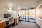 The Arlington 360 Cyber Café is a dedicated business center designed to meet the productivity needs of residents and guests. The space is thoughtfully organized around a series of individual workstations, each equipped with a desktop computer, keyboard, and mouse, set atop clean white countertops with warm wood-toned cabinetry below. Ergonomic rolling chairs in a soft gray upholstery provide comfortable seating at each station, while wall-mounted sconce lighting offers focused illumination over each desk area. A shared printer is conveniently positioned within easy reach, rounding out the practical amenities of the room. The interior is finished with neutral white walls and low-pile carpeting in a subtle pattern, contributing to a calm, focused atmosphere. Access to the room is through a glass-paneled door bearing the Arlington 360 branding, lending the space a professional identity. A framed abstract artwork adorns the corridor wall just outside, adding a touch of curated character to the transition space. Floor-to-ceiling windows nearby flood the surrounding area with natural light and offer views of the landscaped courtyard and neighboring brick residential buildings, connecting the interior environment to the vibrant community outside.