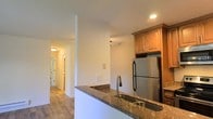 This single-floor space showcases a well-appointed kitchen as its focal point, designed with both functionality and style in mind. The kitchen features dark granite countertops that provide a striking contrast against the warm-toned wood cabinetry above. A stainless steel refrigerator anchors the cooking area, complementing the modern aesthetic of the space. The kitchen includes a double basin sink set into the granite counter, finished with a contemporary arc-neck faucet, adding a practical yet elegant touch.

The layout incorporates a peninsula-style counter that partially separates the kitchen from the adjacent areas, creating a natural flow between spaces. Recessed lighting illuminates the kitchen area, while flush-mount ceiling fixtures extend light throughout the hallway beyond.

Light wood-toned flooring runs consistently throughout the space, lending warmth and visual continuity from the kitchen into the hallway, which leads to additional rooms. The walls are finished in a clean, neutral white, maximizing the sense of brightness and openness. The overall design balances modern finishes with warm, natural tones, creating a cohesive and inviting interior environment.