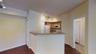 This single-floor residence offers a well-organized and functional layout with a seamless flow between living spaces. The kitchen serves as a central highlight, featuring warm wood cabinetry accented by a decorative wine rack, a full suite of white appliances including a refrigerator, microwave, and range, and a peninsula breakfast bar with a dark countertop that defines the boundary between the kitchen and the adjoining living area. Track lighting installed on the ceiling provides focused illumination over the kitchen workspace, enhancing both functionality and ambiance. The rich, dark hardwood flooring extends throughout the space, adding warmth and visual continuity. Walls finished in a soft, neutral tone create a bright and inviting atmosphere. The layout includes a closet with bi-fold doors, offering practical storage, while doorways leading to additional rooms suggest a thoughtfully designed floor plan. The clean lines, neutral palette, and efficient use of space come together to create a comfortable and livable environment suited for everyday living.