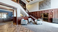The Reserve & Parkway Lounge at 200 E Burnsville Pkwy presents a beautifully appointed community space designed with upscale, transitional aesthetics throughout. The grand lobby greets visitors with soaring ceilings, rich dark wood wainscoting, an intricate herringbone-patterned hardwood floor, and a sweeping staircase with wrought-iron balusters — all accented by contemporary wall art and plush seating arrangements. Adjacent living areas offer multiple gathering spaces, each with its own distinct character. One lounge features a dramatic dark coffered ceiling, large windows with outdoor views, a built-in media wall with cabinetry, and a herringbone-tile fireplace, creating an intimate yet stylish retreat. A secondary lounge continues the refined palette with navy accent walls, a tray ceiling, and mid-century modern furnishings bathed in natural light. The kitchenette area is outfitted with dark wood cabinetry, granite countertops, a navy subway tile backsplash, and pendant lighting over a communal dining table. Perhaps the most distinctive space is the wine lounge, showcasing an arched brick ceiling, stacked stone walls, rich hardwood floors, leather club chairs, and a floor-to-ceiling wine display — an atmosphere of timeless sophistication that ties the entire space together.