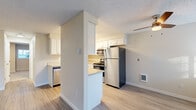 This single-floor residence offers a thoughtfully organized layout with a clean, neutral aesthetic throughout. The kitchen is a functional highlight, featuring white cabinetry, stainless steel appliances including a refrigerator, range, microwave, and dishwasher, complemented by light countertops and wood-style vinyl flooring. A half-wall partition separates the kitchen from the adjacent living area, which benefits from a ceiling fan and direct access to the outdoors through sliding glass doors that invite natural light and views of surrounding greenery. Additional rooms include carpeted spaces well-suited for bedrooms, with one featuring a window with blinds overlooking the exterior. The bathroom is neatly designed with a full tub-shower surround, a single-sink vanity with white cabinetry, and wood-look tile flooring. Warm beige tones on the walls carry consistently throughout the unit, creating a cohesive and inviting atmosphere. Textured ceilings are present in all rooms, and wall-mounted heating units provide climate control. The overall layout is efficient and practical, offering comfortable living spaces with a straightforward, move-in-ready presentation.