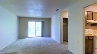 This single-floor space features a spacious, open main room finished with wall-to-wall carpeting in a neutral tone, providing a comfortable and cohesive foundation throughout. The room is anchored by a large sliding glass door that fills the space with natural light and offers direct access to an outdoor area, where mature greenery is visible beyond the railing. The textured popcorn ceiling adds a classic character overhead, while a central HVAC vent ensures year-round climate control. Neutral-toned walls throughout the main room create a clean, versatile backdrop adaptable to a variety of interior styles. Adjacent to the main room, an open closet alcove provides practical built-in storage, featuring double hanging rods that maximize vertical storage capacity. The closet's open design makes for easy access and organization. White baseboards and door trim provide clean architectural contrast against the warm wall tones. Overall, the space presents a straightforward, functional layout with an emphasis on natural light, storage, and comfortable living in a well-maintained, move-in-ready condition.