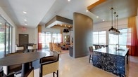 This single-floor space is a thoughtfully designed communal area that seamlessly blends lounge, dining, and bar functions within an open-plan layout. The interior is defined by striking architectural details, including a curved accent column, layered tray ceilings with recessed lighting, and bold orange accent beams that add a contemporary edge to the design.

The lounge area is anchored by a comfortable seating arrangement with patterned upholstered sofas and armchairs set atop a warm area rug, centered around a coffee table. Large floor-to-ceiling windows dressed with sheer and semi-opaque curtains in warm red and orange tones flood the space with natural light and offer outward views.

Adjacent to the lounge, a bar counter finished in richly veined dark granite provides a sophisticated social focal point, paired with woven high-back bar stools. A pendant light with a bold black and gold finish hangs overhead, adding a decorative statement.

The dining area features a round dark wood table with woven-back chairs, complementing the overall warm, contemporary color palette. The polished light-toned flooring runs throughout, tying the distinct zones together cohesively. The overall ambiance is welcoming, energetic, and well-appointed, making this a functional and visually engaging communal gathering space.