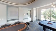 Located at Johnson and Wales Way in Charlotte, NC, this single-floor amenity lounge is designed to offer a dynamic and social environment. The space is defined by its open layout, patterned carpet in blue and gray tones, and an abundance of natural light streaming through floor-to-ceiling windows and large glass panels that frame views of surrounding greenery and the urban streetscape beyond.

A bold blue accent wall serves as a focal point, housing a wall-mounted flat-screen television and providing a vibrant contrast to the otherwise neutral gray palette throughout. The game area is well-appointed, featuring a poker table with surrounding seating, a billiards table, and a foosball table, making it a versatile hub for recreation and entertainment.

A wet bar area along one wall includes dark wood cabinetry, a countertop with a sink, and high-top bistro tables positioned beside windows overlooking an outdoor pool area. Cylindrical columns and a curved tray ceiling detail add architectural interest to the space. Elevator access and tiled entryway corridors connect the lounge to the broader building, while decorative wall art adds a finishing touch to the contemporary interior.