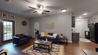 This single-story unit offers a cohesive, modern interior defined by neutral gray walls, wood-style flooring, and white trim that carry consistently through every room. The living area serves as the central hub of the space, accommodating a comfortable seating arrangement beneath a ceiling fan with light fixture. A front entry door provides natural light into the room, and the open sightlines connect the living area to the kitchen, which features dark cabinetry, granite-style countertops, black appliances including a range and over-the-range microwave, and a pantry with a six-panel door.

Two bedrooms are accessible from the main living area. The larger bedroom includes a ceiling fan, ample floor space, and a walk-in closet. The second bedroom is well-suited as a guest room or home office, featuring a window with blinds that allows natural light to fill the room. Both bedrooms share access to two full bathrooms, each outfitted with a dark wood-tone vanity, granite-style countertop, undermount sink, a full mirror, and a tub-shower combination. The bathrooms continue the wood-style flooring seen throughout the unit, reinforcing the space's consistent and practical design aesthetic.