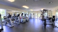 This single-floor fitness center offers a functional and well-organized workout environment designed to accommodate a range of exercise needs. The space is outfitted with multiple elliptical trainers and stationary bikes arranged in a row, providing ample cardio options for users. A multi-station strength training machine is positioned to one side, offering resistance-based exercise opportunities. A full-length mirror spans one wall, enhancing the sense of space while allowing users to monitor their form during workouts. The flooring consists of dark rubberized material, providing durability, impact absorption, and safety during exercise. Overhead fluorescent lighting ensures the space is well-lit throughout, supplemented by natural light streaming through multiple windows and glass-paneled doors. The doors lead directly to an outdoor area, promoting ventilation and easy access. The walls are finished in a neutral light tone, contributing to a clean and open atmosphere. The overall layout is practical and spacious, with adequate room between equipment to allow comfortable movement. The facility presents a straightforward, no-frills environment focused on providing residents or members with accessible fitness amenities.