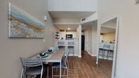 This two-story residence offers a well-designed layout that balances functionality and comfort across both levels. The main floor features an open-concept living and dining area anchored by warm hardwood-style flooring that extends throughout the home. A large window in the living room frames outdoor views and fills the space with natural light. The adjacent dining area accommodates a full table with seating, while the kitchen is equipped with white cabinetry, a double-basin sink, a dishwasher, and black appliances including a range and built-in microwave. A breakfast bar with counter seating connects the kitchen to the dining space. The upper level opens to a versatile loft-style common area with wall-mounted shelving, ideal for a study or lounge. Two well-proportioned bedrooms are found on this floor, each featuring hardwood-style flooring, large windows that bring in natural light, and dedicated desk areas. Both bathrooms include a tub-shower combination, white vanity cabinetry with countertop sinks, and hardwood-style flooring. A generously sized walk-in closet with wire shelving and hanging rods provides ample storage. Neutral tones and clean finishes give the entire space a cohesive, contemporary feel.