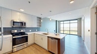 Located at 601 Bond Ave in Grand Rapids, MI, this well-appointed one-bedroom, one-bathroom unit offers a thoughtfully designed open layout with contemporary finishes throughout. The combined kitchen and living area serves as the heart of the home, featuring an open-concept design with a peninsula island topped with white quartz countertops, white flat-panel cabinetry, a blue-gray tile backsplash, and a full suite of stainless steel appliances including a refrigerator, electric range, microwave, and dishwasher. Pendant lighting above the island and warm wood-tone flooring add a refined, modern touch. The living area is anchored by dramatic floor-to-ceiling windows that frame sweeping views of the Grand River, downtown Grand Rapids skyline, and surrounding greenery, flooding the space with natural light. The bedroom is a quiet, comfortable retreat with its own large window capturing additional city views and serene natural light. A sliding barn door opens to reveal a closet with built-in shelving alongside a stacked in-unit washer and dryer. The bathroom is finished with large-format tile in the tub/shower surround, a white vanity with ample drawer storage, and open wall shelving for added functionality.