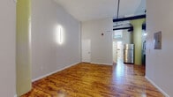 This single-floor loft-style unit masterfully combines industrial character with contemporary finishes, creating a space that feels both open and inviting. Rich, warm-toned hardwood floors run throughout the main living areas, reflecting natural light and adding a sense of depth and warmth to the space.

The main living area is generously proportioned, with soaring ceilings that amplify the airy, open atmosphere. Sleek vertical wall-mounted light fixtures provide modern accent lighting, while exposed structural beams and a distinctive column add authentic loft character. A bedroom features large, black-framed windows set against a painted brick wall, flooding the room with natural light and offering urban rooftop views.

The kitchen is well-appointed with dark cabinetry, white quartz countertops, subway tile backsplash, stainless steel appliances including a gas range and dishwasher, and open wood shelving with black metal brackets — a stylish mix of industrial and contemporary design.

The bathroom is finished with clean white subway tile surrounds in the tub/shower, a dark wood vanity with a white basin, matte black fixtures throughout, and a round mirror, delivering a modern and cohesive aesthetic. The overall ambiance of this space speaks to urban loft living with thoughtfully curated modern updates.