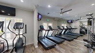 Located at 1919 Post Oak Park Dr in Houston, TX, this well-appointed fitness center offers a comprehensive workout environment within a compact, efficiently designed single-floor layout. The exercise room features wood-look flooring throughout, lending a clean and modern aesthetic that is both practical and visually appealing.

The cardio zone is prominently equipped with a row of treadmills, elliptical trainers, a stair climber, and stationary spin bikes, providing a wide range of aerobic options. For strength training, the space includes a cable machine, a multi-functional weight station, a full rack of dumbbells in graduated sizes, and an adjustable bench, catering to a variety of workout routines.

Large wall-to-wall mirrors visually expand the room and allow users to monitor their form, while multiple wall-mounted flat-screen TVs offer entertainment during workouts. Dual ceiling fans contribute to air circulation and comfort throughout the space. Natural light streams in through several windows and glass-paneled doors, creating a bright and energizing atmosphere.

Crown molding and decorative columns add subtle architectural character to the room, elevating the overall design. Recessed lighting supplements natural light during evening hours. The result is a polished, fully functional fitness center that balances form and function for an effective workout experience.