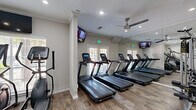 Located at 1919 Post Oak Park Dr in Houston, TX, this well-appointed fitness center offers a comprehensive workout environment within a compact, efficiently designed single-floor layout. The exercise room features wood-look flooring throughout, lending a clean and modern aesthetic that is both practical and visually appealing.

The cardio zone is prominently equipped with a row of treadmills, elliptical trainers, a stair climber, and stationary spin bikes, providing a wide range of aerobic options. For strength training, the space includes a cable machine, a multi-functional weight station, a full rack of dumbbells in graduated sizes, and an adjustable bench, catering to a variety of workout routines.

Large wall-to-wall mirrors visually expand the room and allow users to monitor their form, while multiple wall-mounted flat-screen TVs offer entertainment during workouts. Dual ceiling fans contribute to air circulation and comfort throughout the space. Natural light streams in through several windows and glass-paneled doors, creating a bright and energizing atmosphere.

Crown molding and decorative columns add subtle architectural character to the room, elevating the overall design. Recessed lighting supplements natural light during evening hours. The result is a polished, fully functional fitness center that balances form and function for an effective workout experience.