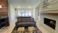This single-floor space is thoughtfully designed for both relaxation and entertainment, blending modern aesthetics with natural, rustic accents. The open layout centers around a full-size pool table positioned beneath a bank of large windows that frame sweeping views of mature trees, filling the room with abundant natural light. A charcoal tufted sofa anchors the lounge area, complemented by unique live-edge wood coffee tables that add an organic, earthy touch to the contemporary setting. The flooring throughout features light-toned tile that enhances the bright, airy atmosphere of the space. On one end of the room, a striking stone fireplace serves as a dramatic focal point, with a reclaimed wood mantel adding warmth and texture to the clean white walls. Recessed lighting overhead ensures the space is well-lit regardless of the time of day. Bar-height stools along the window wall provide additional casual seating, making this a versatile gathering area. The overall ambiance balances a relaxed, inviting feel with refined design details, making it well-suited for both casual hangouts and entertaining guests.
