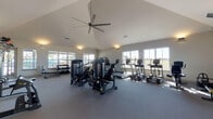 This fitness center offers a thoughtfully designed, single-floor workout facility that combines functionality with a bright, open atmosphere. The space is defined by its vaulted ceilings, which lend an airy, expansive feel throughout, while a large ceiling fan ensures comfortable air circulation during workouts. Multiple windows line the perimeter walls, flooding the interior with natural light and providing views of the surrounding outdoor areas, including what appears to be a pool and recreational space.

The main workout floor is finished with durable, gray fitness flooring and is divided into dedicated zones for various training modalities. The strength training area features a comprehensive selection of cable machines, weight machines, and a dumbbell rack with a flat bench, catering to a full range of resistance exercises. Full-length mirrors are mounted along one wall, enhancing the sense of space and allowing users to monitor their form. A wall-mounted flat-screen TV is positioned for easy viewing from this area.

The cardio zone is well-stocked with a row of True-brand treadmills, elliptical trainers, and additional cardio machines. A separate, open stretching and functional training room provides space for floor-based exercises, battle ropes, and ab equipment, also equipped with a wall-mounted TV and large windows. A water fountain station is conveniently accessible near the entrance, and a rustic sliding barn door leads to an adjacent outdoor area.