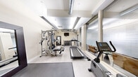 Located at 770 N Grant St in Denver, Colorado, this fitness center offers a well-equipped exercise space designed to support a full range of workout routines. The room features light-toned walls and a neutral tile floor, creating a clean and functional environment. Large floor-to-ceiling windows with horizontal blinds line one side of the space, allowing ample natural light to filter in while maintaining privacy.

The exercise room is outfitted with a variety of equipment to accommodate both cardiovascular and strength training. Cardio options include a Spirit treadmill and a recumbent exercise bike, each positioned on protective floor mats. A multi-station cable machine and a Nautilus functional trainer provide comprehensive strength training capabilities, complemented by a dedicated free weight area featuring a tiered dumbbell rack with a full range of weights. A flat workout bench and stability balls round out the available training tools. A wall-mounted flat-screen television is positioned for easy viewing during workouts.

Exposed ductwork along the ceiling adds an industrial character to the otherwise clean, modern aesthetic. A full-length mirror with a dark frame enhances the sense of space and supports form-checking during exercise. The room is well-lit with recessed and linear ceiling fixtures, and includes clearly marked exits for safety.