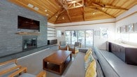 The Upper Pool Clubhouse is a thoughtfully designed single-floor community gathering space that blends natural materials with modern aesthetics. The most striking architectural feature is the soaring vaulted ceiling, clad entirely in warm-toned wood planks and supported by exposed timber trusses, lending the space a refined yet welcoming character. Recessed lighting and a decorative branch chandelier complement the natural wood overhead.

The main lounge area is anchored by a floor-to-ceiling stone fireplace finished in stacked grey slate tile, with a wall-mounted flat-screen television above a wood mantel. Built-in shelving and cabinetry flank the fireplace, providing both display and storage space. Comfortable grey seating with colorful accent pillows and solid wood-framed chairs are arranged around a substantial wood coffee table, creating an inviting social setting.

Polished concrete floors run throughout, offering a clean, contemporary contrast to the warmth of the wood ceiling. Large glass sliding and French doors on multiple walls flood the interior with natural light and provide views of the surrounding outdoor pool area and landscaping. A wooden console table with task lighting adds a functional workspace element along one wall. Framed artwork throughout contributes to the polished, curated ambiance of this community clubhouse.