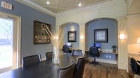 This well-appointed single-floor space blends classic architectural elegance with modern functionality. The interior is immediately distinguished by its striking arched alcoves framed with crisp white wainscoting and molding, which contrast beautifully against the cool blue-gray walls. Each alcove houses a dedicated computer workstation complete with granite-topped desks, ergonomic task chairs, and framed wall art, making them ideal for focused individual work or research.

The central area features a substantial dark wood conference or meeting table surrounded by leather upholstered chairs, providing an inviting setting for collaboration or group gatherings. A tall glass vase with decorative branches serves as an elegant centerpiece, adding a touch of organic warmth to the space.

Overhead, a coffered ceiling with recessed lighting and track lighting ensures the space is well-illuminated throughout. Rich hardwood flooring runs the length of the room, adding warmth and a sense of continuity. Wall-mounted sconces within the alcoves provide soft, ambient lighting that complements the overall atmosphere.

The space successfully combines a professional, business-ready environment with refined interior design details, resulting in an atmosphere that is both functional and visually sophisticated.