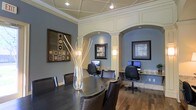 This well-appointed single-floor space blends classic architectural elegance with modern functionality. The interior is immediately distinguished by its striking arched alcoves framed with crisp white wainscoting and molding, which contrast beautifully against the cool blue-gray walls. Each alcove houses a dedicated computer workstation complete with granite-topped desks, ergonomic task chairs, and framed wall art, making them ideal for focused individual work or research.

The central area features a substantial dark wood conference or meeting table surrounded by leather upholstered chairs, providing an inviting setting for collaboration or group gatherings. A tall glass vase with decorative branches serves as an elegant centerpiece, adding a touch of organic warmth to the space.

Overhead, a coffered ceiling with recessed lighting and track lighting ensures the space is well-illuminated throughout. Rich hardwood flooring runs the length of the room, adding warmth and a sense of continuity. Wall-mounted sconces within the alcoves provide soft, ambient lighting that complements the overall atmosphere.

The space successfully combines a professional, business-ready environment with refined interior design details, resulting in an atmosphere that is both functional and visually sophisticated.