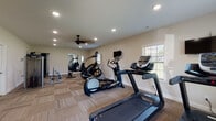 This fitness center is a thoughtfully designed, single-floor workout facility built to support a full range of exercise routines. The open layout provides ample space for movement between stations, with patterned carpet tile flooring throughout that adds comfort underfoot during workouts.

The cardio section is prominently featured and includes multiple treadmills, an elliptical trainer, and a stair climber, offering a variety of options for aerobic exercise. For strength training, the facility includes a Precor functional trainer cable machine, a multi-station weight machine, and free weight equipment, enabling both resistance and full-body conditioning workouts.

A large wall mirror spans one end of the room, enhancing the sense of space while allowing users to monitor their form. A wall-mounted flat-screen TV provides entertainment during cardio sessions. The ceiling fan ensures comfortable air circulation, complementing the recessed lighting that keeps the space bright and well-lit.

Natural light enters through several windows with blinds, offering views of the surrounding greenery. Practical amenities include a sanitation station with paper towels and spray cleaner, a waste bin, and a fire extinguisher. A dedicated exit door provides convenient access directly to the outdoors, making this fitness center a functional and inviting space for daily exercise.