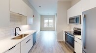 Located at 845 Greenfield Rd in Lancaster, PA, this thoughtfully designed single-floor unit offers a clean, contemporary aesthetic throughout its efficiently utilized layout. The kitchen serves as a functional centerpiece, featuring flat-front white cabinetry extending to the upper walls, a white quartz countertop, a subway tile backsplash, and a suite of stainless steel appliances including a range, built-in microwave, dishwasher, and refrigerator. A single-basin sink with a matte black faucet adds a modern finishing touch. Light hardwood-style flooring runs continuously throughout the space, lending warmth and visual cohesion. The open living area beyond the kitchen benefits from natural light streaming through a large window, creating a bright and airy atmosphere. A ceiling fan with a sleek black finish provides both function and style in the bedroom area, which also features a window overlooking the exterior. Additional rooms present clean, neutral walls with generous ceiling height, white panel doors with black hardware, and ample room for flexible furnishing arrangements. The overall design embraces a minimalist, move-in-ready aesthetic with consistent finishes that give the space a polished, cohesive feel.